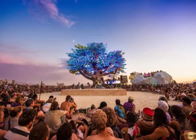 De interactieve lichtboom in paars en blauwe kleuren staat in het midden van een cirkel, omringd door tientallen mensen op het festival Burning Man, met een prachtige blauw paarse lucht op de achtergrond.